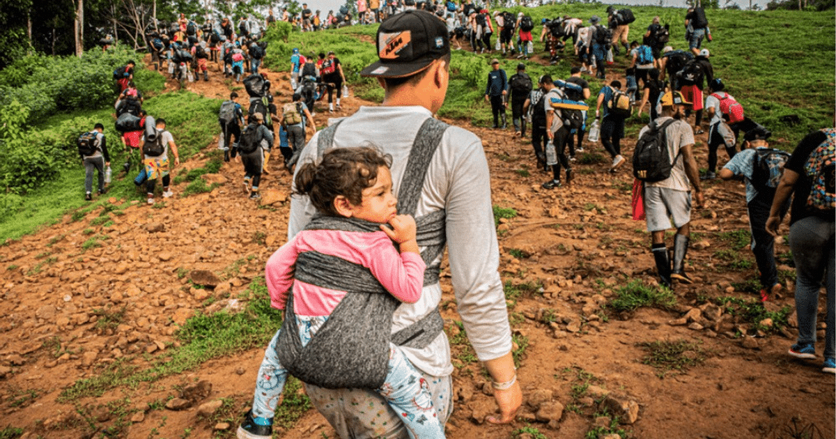 Grupo de personas migrando por un sendero rocoso, liderado por un adulto con una niña en la espalda.