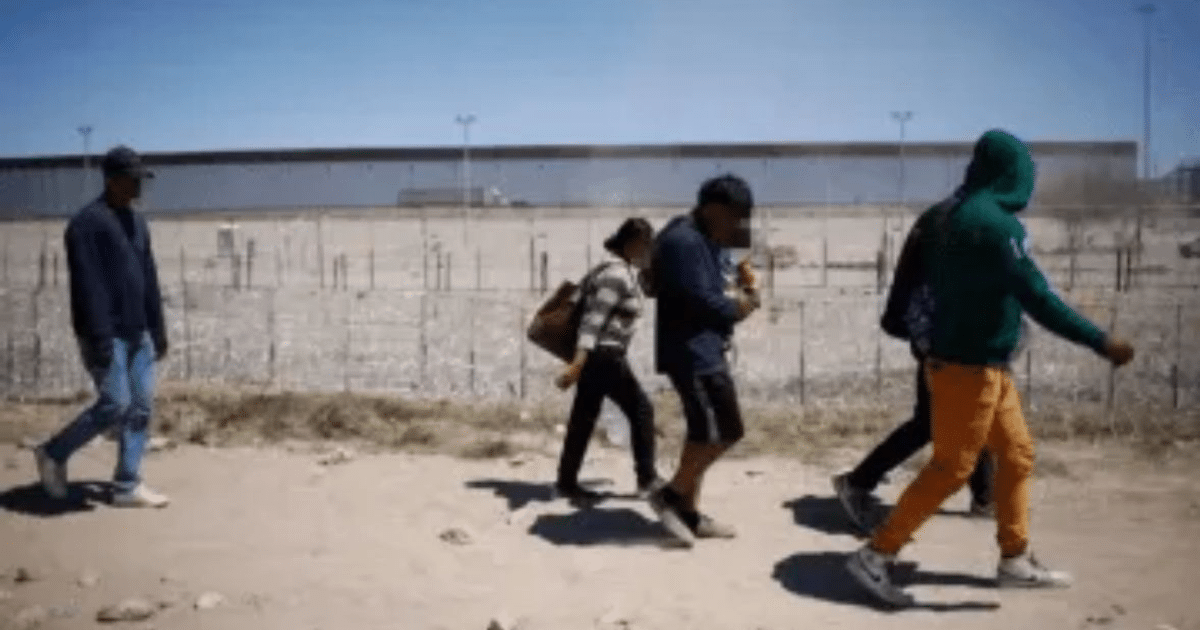 Grupo de migrantes caminando en un desierto con una valla al fondo, simbolizando la migración en la frontera.