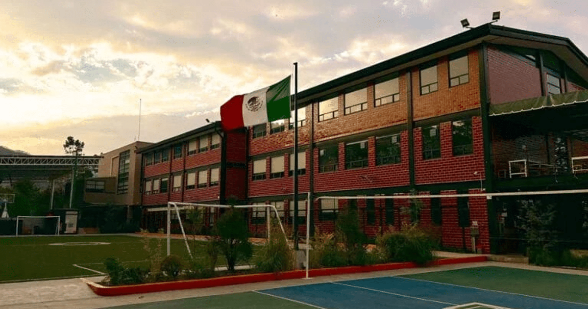 Edificio escolar de ladrillos en Sinaloa con cancha de fútbol y bandera mexicana al atardecer.