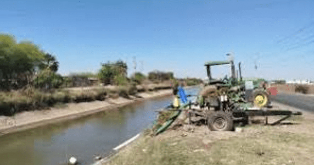 Canal de agua en Ahome con tractor verde y vegetación, día soleado.