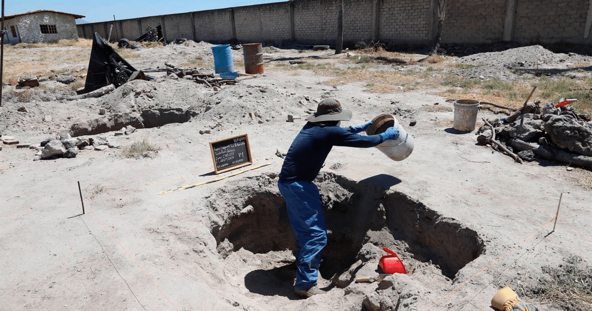 Trabajador en excavación árida con sombrero y balde, rodeado de herramientas y barriles.