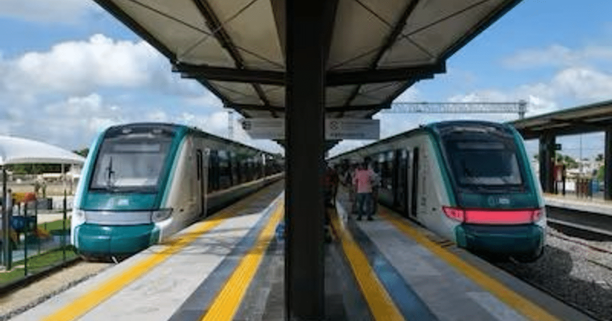 Dos trenes de cercanías en una estación moderna con personas abordando, cielo despejado y área verde al fondo.