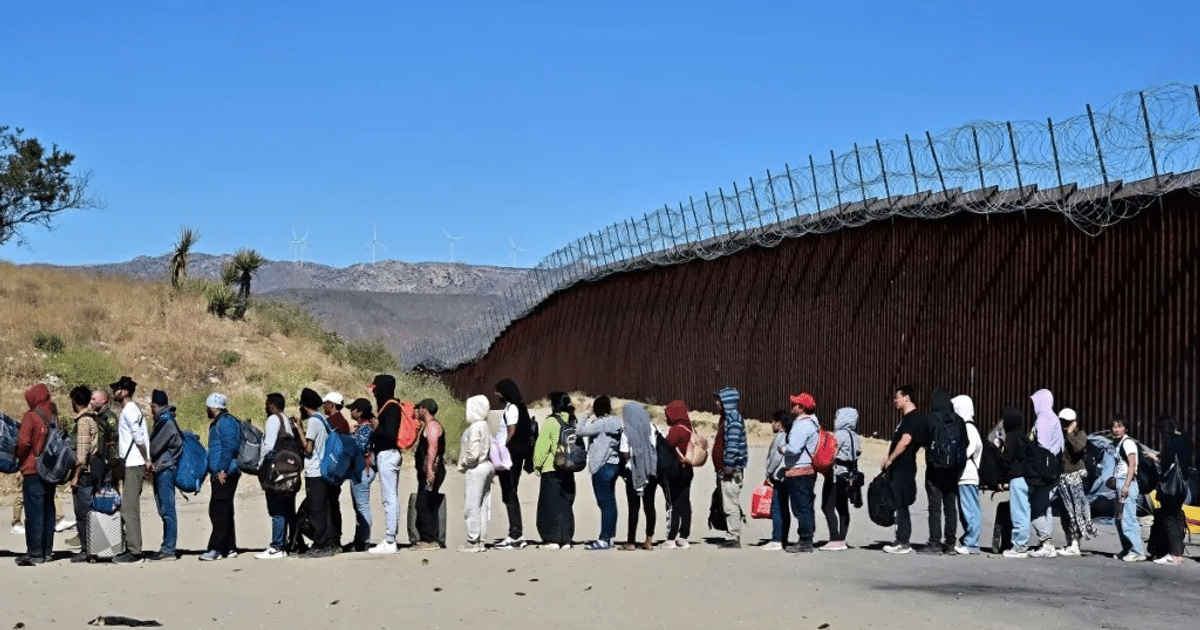 Grupo de personas con mochilas esperando frente a una valla, con un parque eólico al fondo.