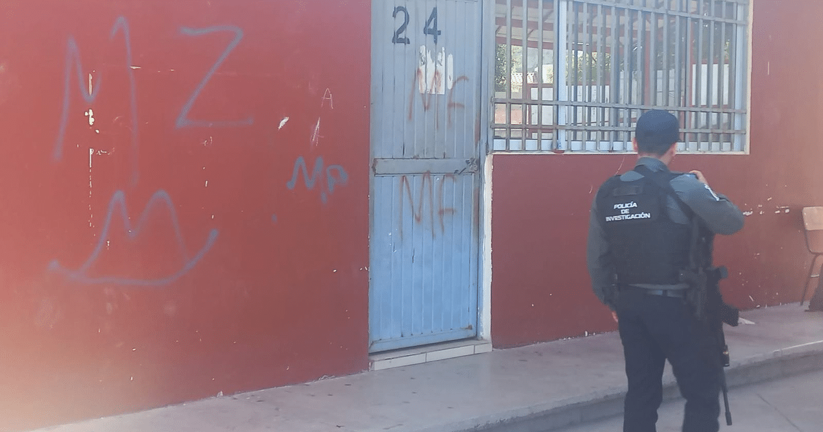 Oficial de policía investigando grafitis en una pared roja junto a una puerta azul en Culiacán.