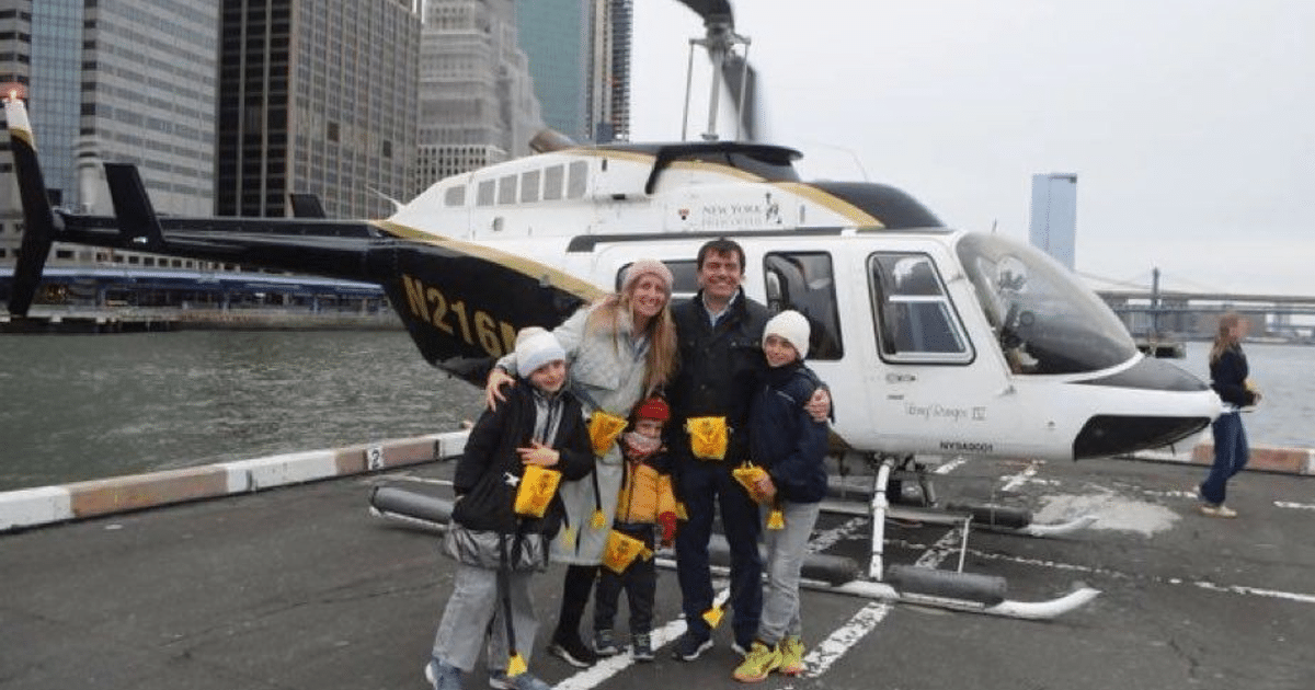 Familia con bolsas amarillas junto a un helicóptero en un puerto, rascacielos al fondo.