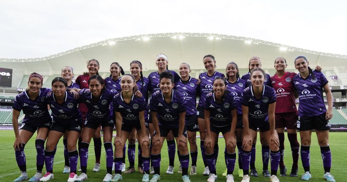 Jugadoras de fútbol femenil posando en un campo, con camisetas moradas y una portera en camiseta roja, en un evento de presentación del equipo.