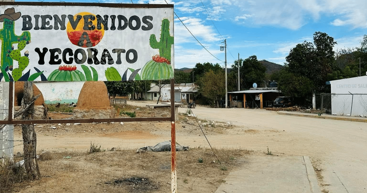 Señal de bienvenida a Yecorato con cactus y flores, carretera de tierra y montañas al fondo.
