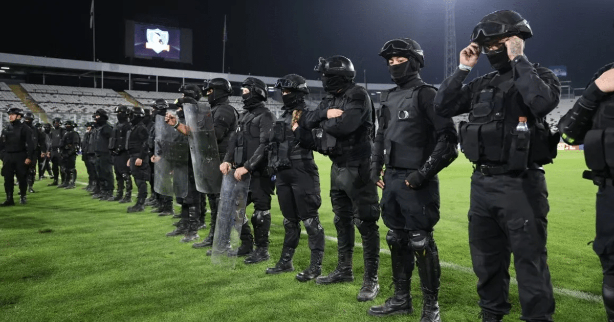 Fuerzas de seguridad alineadas en el Estadio Monumental, equipadas con escudos y atuendos de protección, en un evento deportivo.