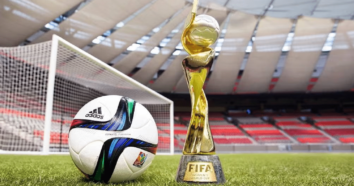 Balón Adidas y trofeo de la Copa Mundial Femenina en campo de fútbol con portería y gradas al fondo.