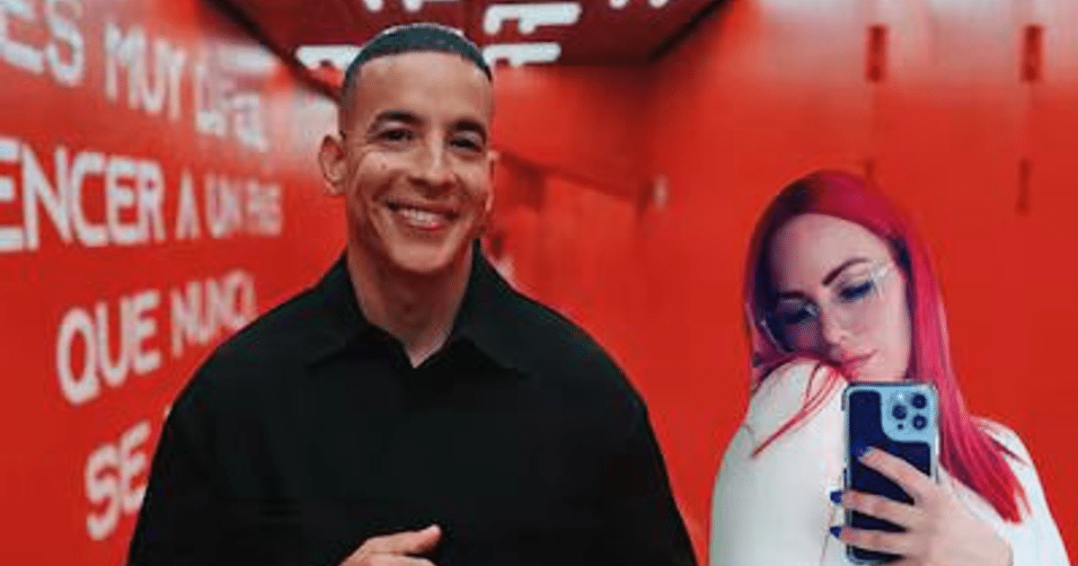 Dos personas en un fondo rojo, una sonríe y la otra toma una selfie, con texto en la pared detrás.