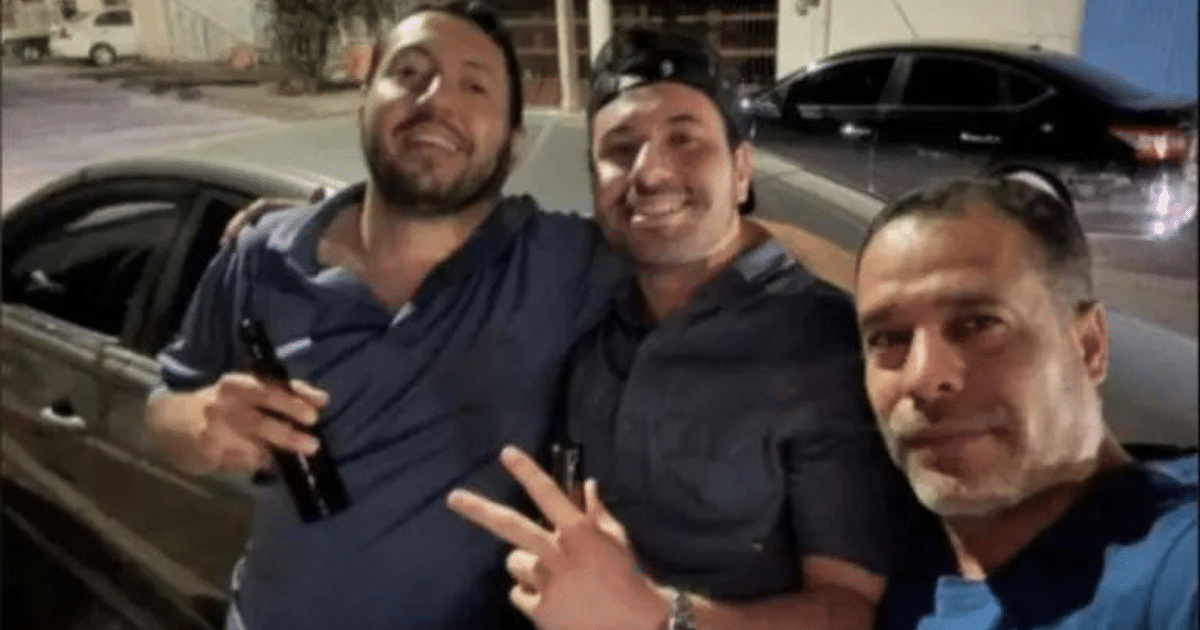 Tres hombres sonrientes posando juntos en la noche con un automóvil al fondo.