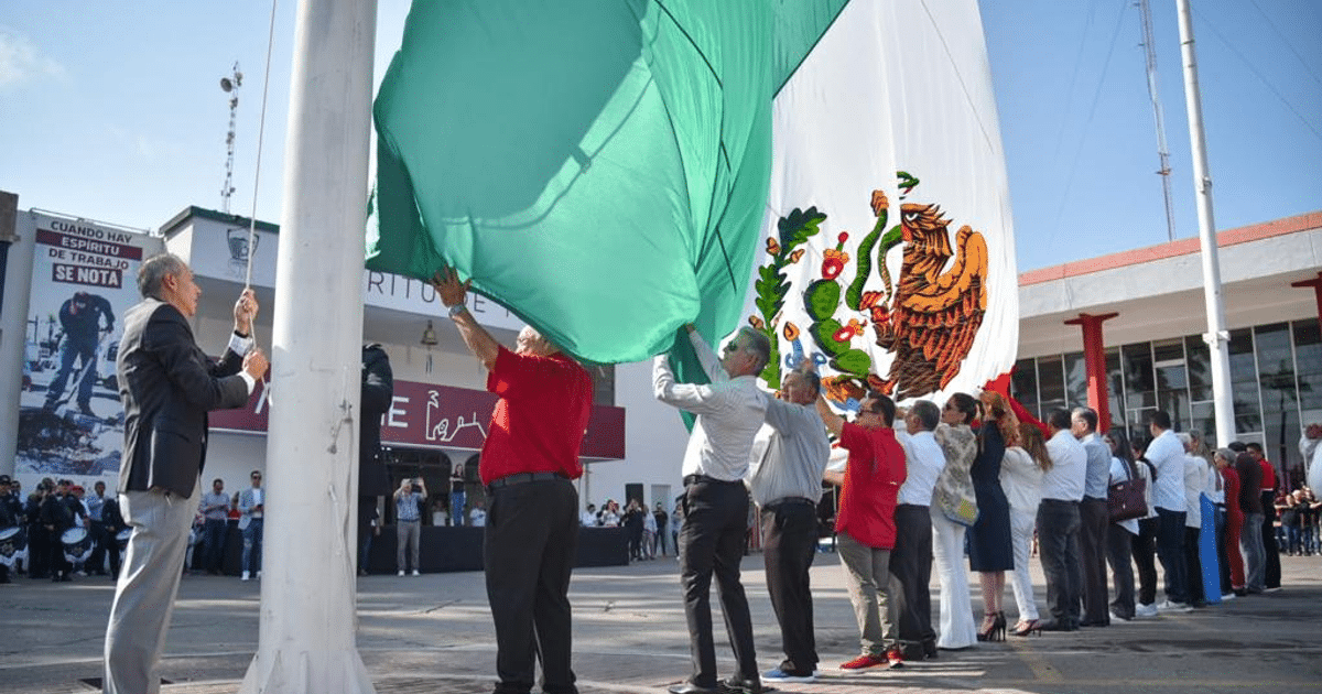 Personas izando la bandera de México en un acto cívico en Los Mochis, con un edificio al fondo que tiene un mensaje sobre el trabajo en equipo.