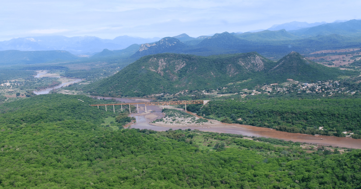 Paisaje natural con montañas verdes, río y puente en Choix, reflejando tranquilidad en medio de tensiones.
