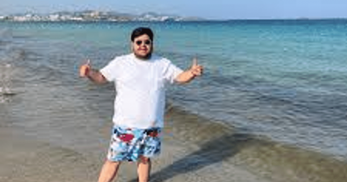 Persona en la playa con camiseta blanca y bañador colorido, haciendo gesto positivo con el mar de fondo.