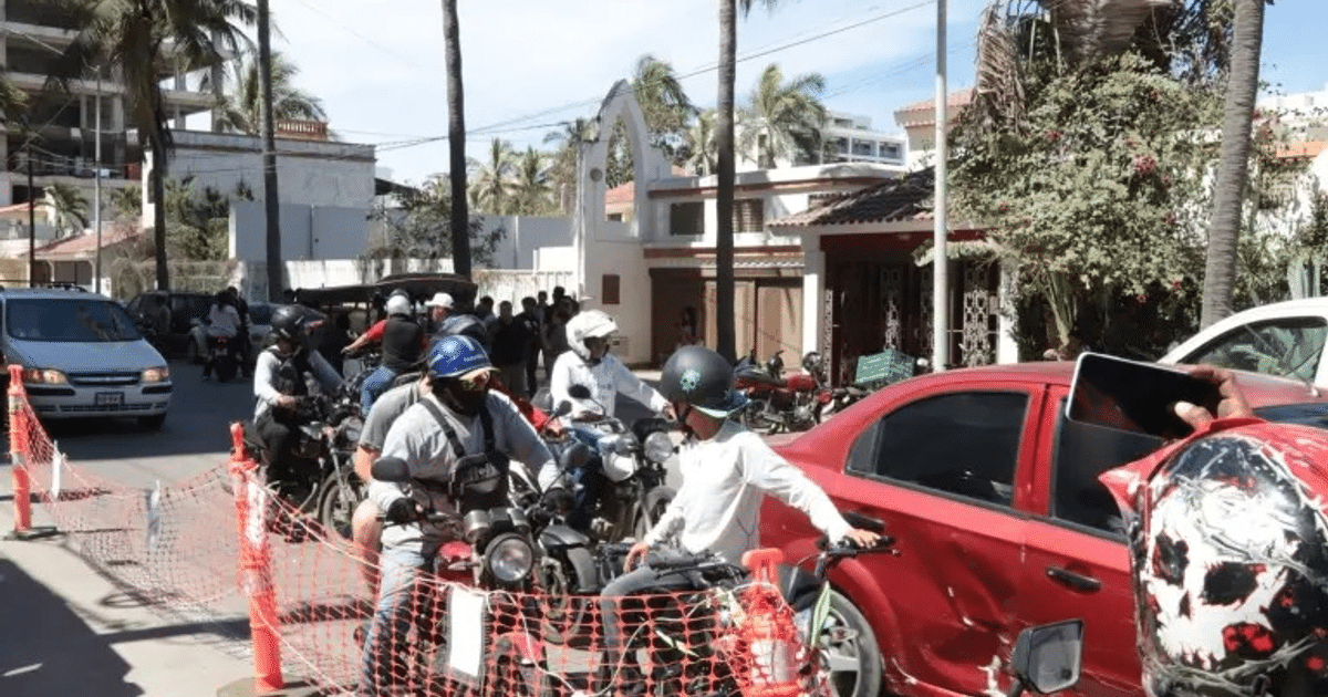 Calle de Mazatlán con tráfico, motocicletas, vehículos y personas usando cascos, con árboles y edificios al fondo.