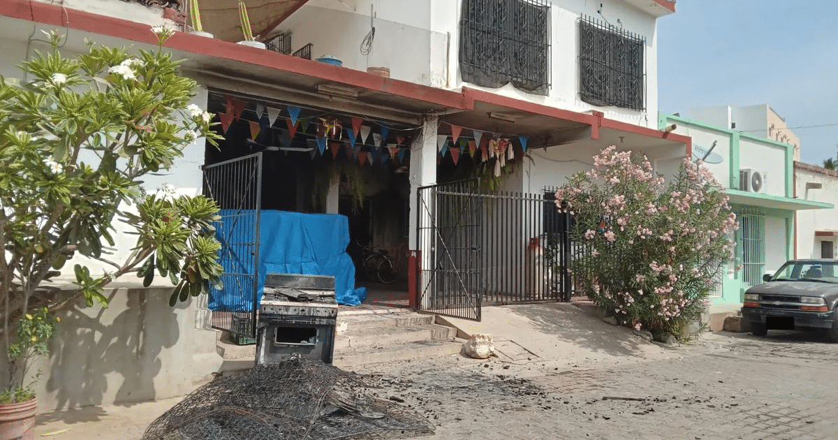 Fachada de edificio urbano con puerta metálica, toldo azul, plantas, vehículo y escombros, decorada con banderines de colores.