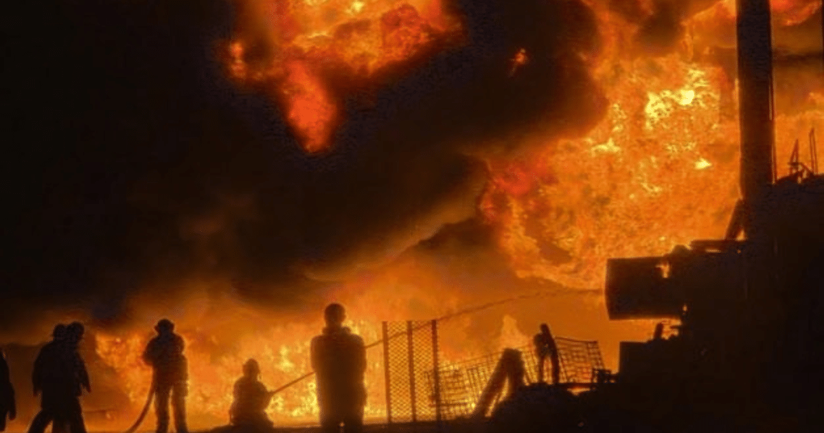 Escena de incendio en almacén de combustible con llamas y humo, y siluetas de personas respondiendo al siniestro.