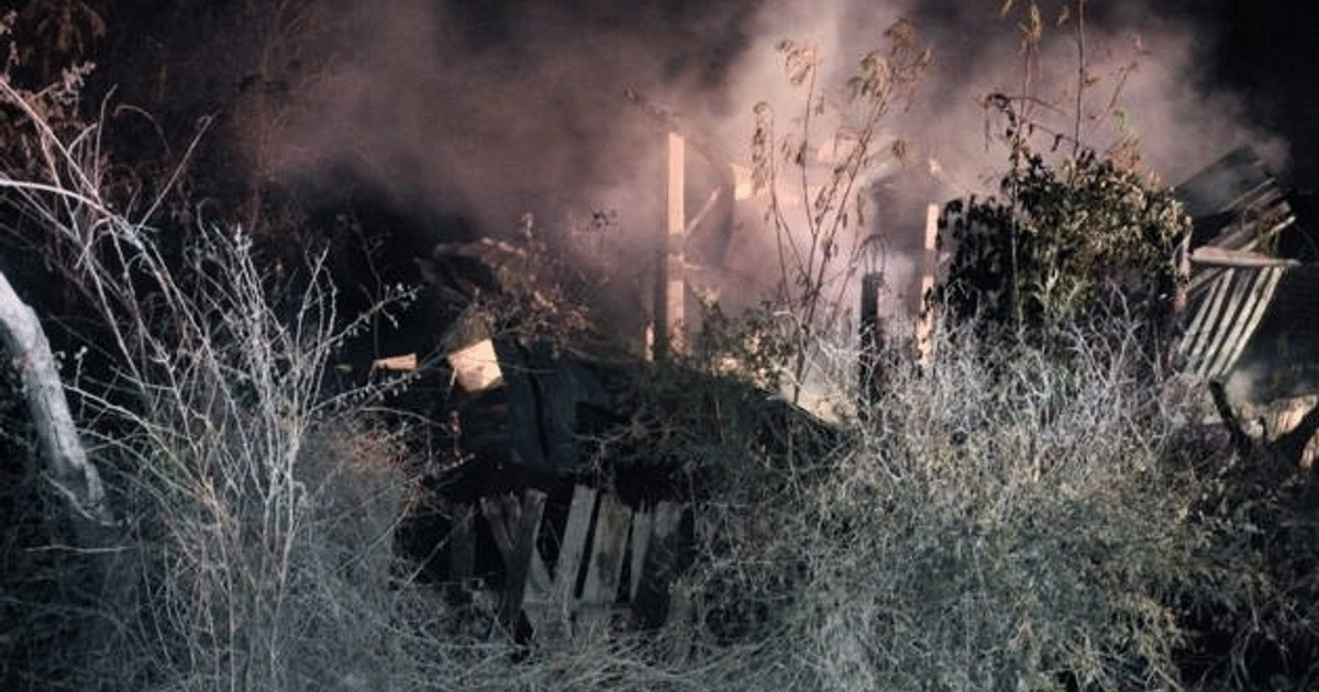 Restos de una estructura quemada rodeada de humo y maleza seca en un entorno nocturno.