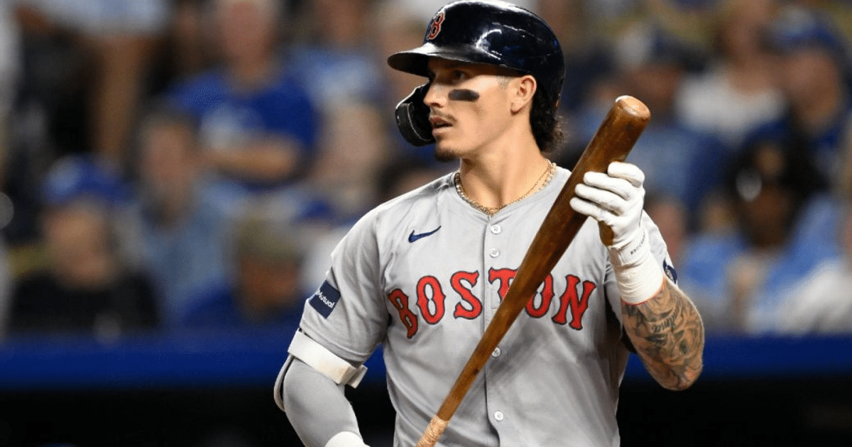 Jugador de béisbol de Boston con uniforme gris y bate en el estadio, con aficionados al fondo.