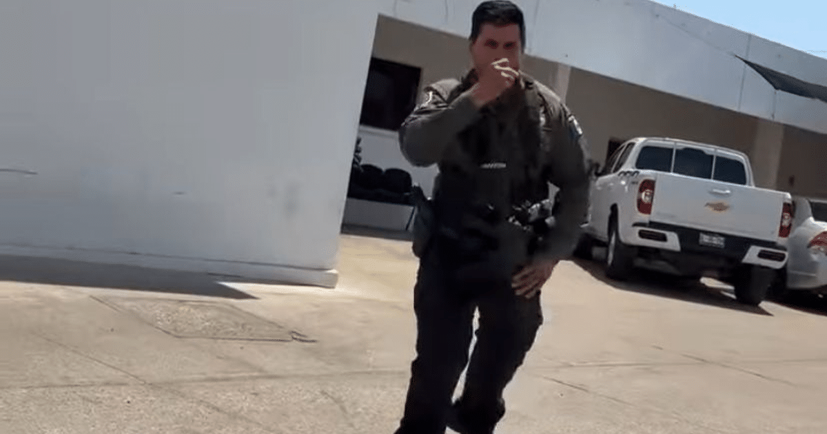 Agente de policía en uniforme acercándose a la cámara en un entorno al aire libre con vehículos estacionados y una estructura blanca de fondo.