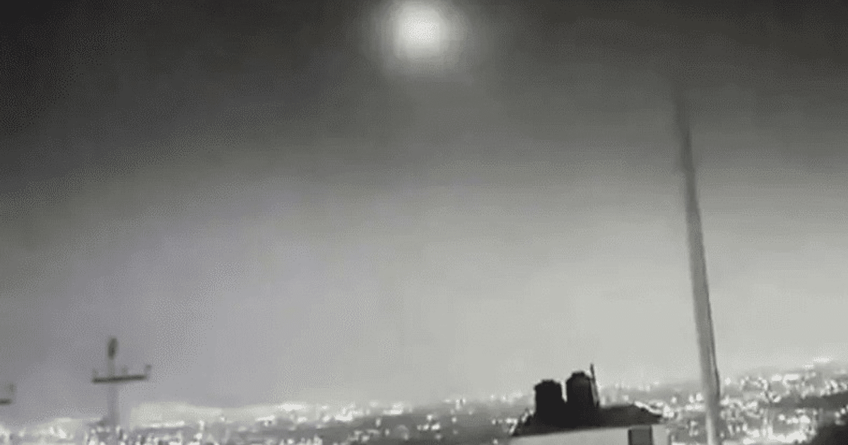 Escena nocturna en blanco y negro con un objeto brillante en el cielo y luces de ciudad en el fondo.