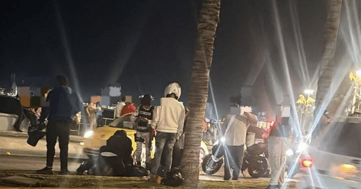 Grupo de personas en una calle nocturna de Mazatlán con un coche amarillo y palmeras, relacionado con un accidente vial reciente.