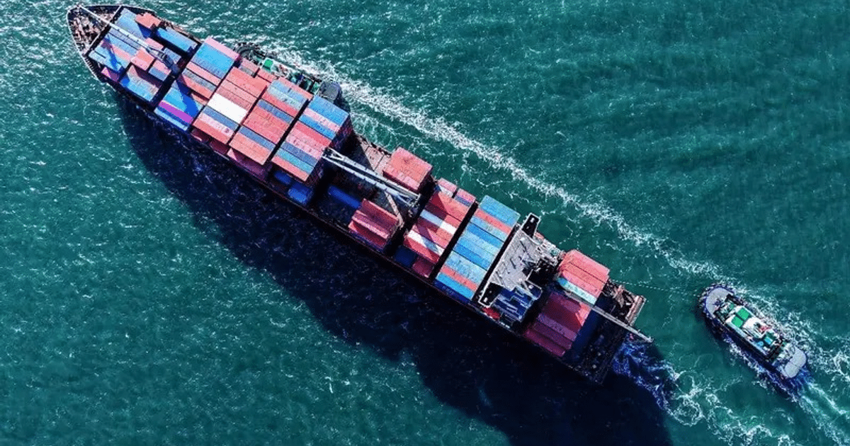 Barco de carga con contenedores multicolores y remolcador en aguas azules, visto desde arriba.