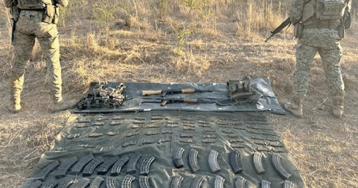 Dos personas con ropa militar junto a armas y cargadores en una zona rural.