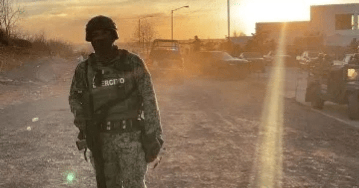 Soldado en uniforme táctico de pie en una carretera polvorienta al atardecer, con vehículos al fondo.