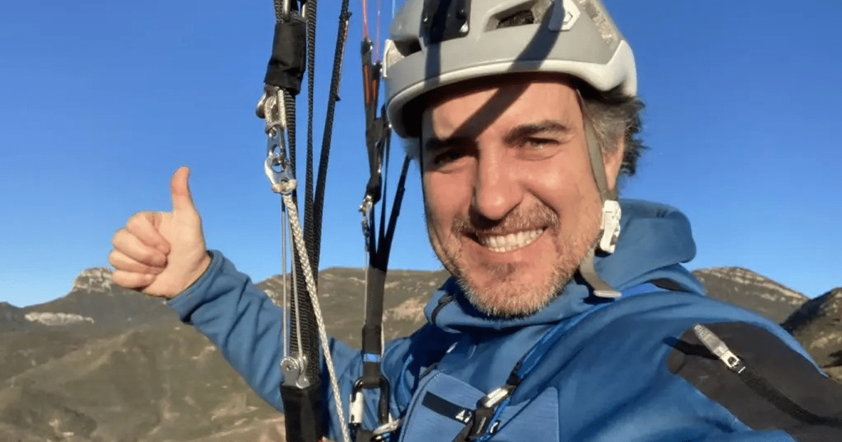 Persona con casco y chaqueta azul sonriendo y sosteniendo cuerdas para parapente, con montañas al fondo.