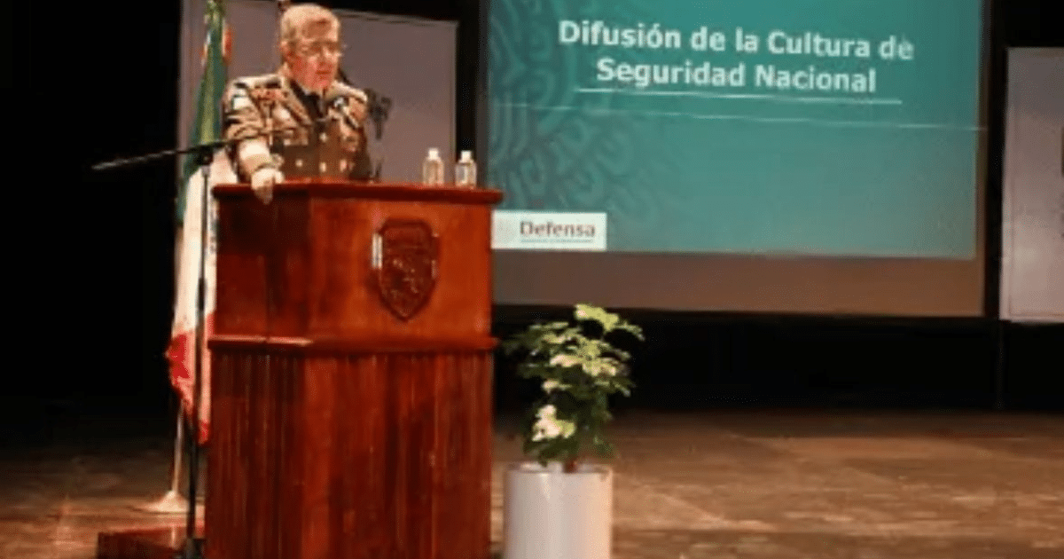 Persona dando un discurso sobre seguridad nacional en la UAS con bandera mexicana de fondo.