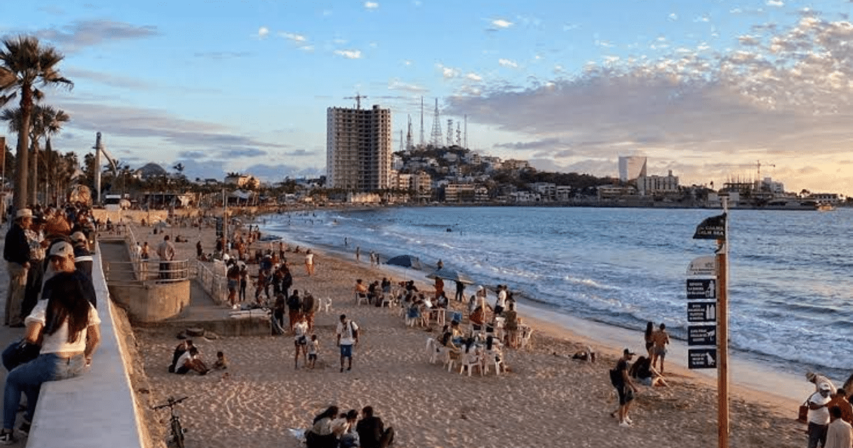 Playa de Mazatlán con turistas, malecón y edificios al fondo en un día soleado.