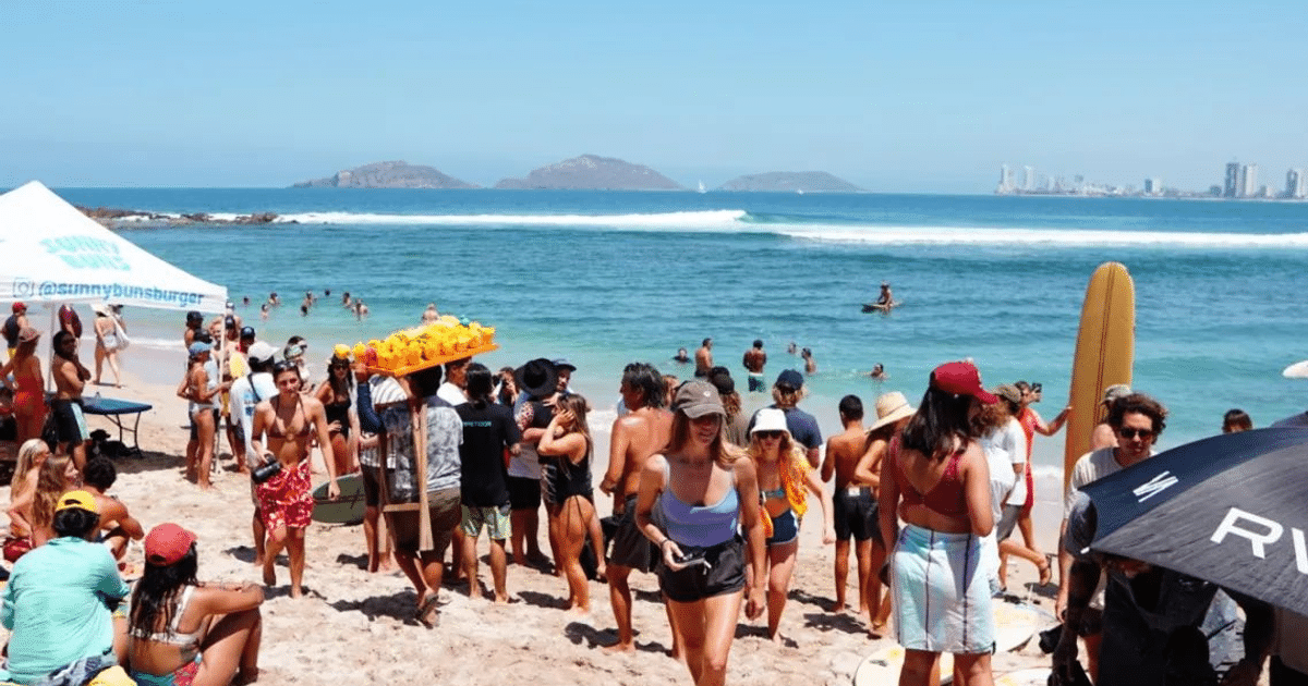 Playa concurrida en Mazatlán con personas disfrutando del sol, surfistas en el agua y un vendedor ambulante.