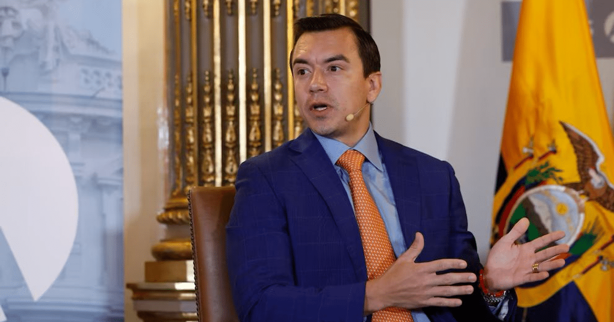 Hombre en traje azul con corbata naranja discutiendo en evento formal con bandera de Ecuador de fondo