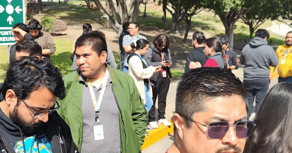 Grupo de personas reunidas al aire libre con teléfonos, árboles y letrero de punto de reunión al fondo en un día soleado.