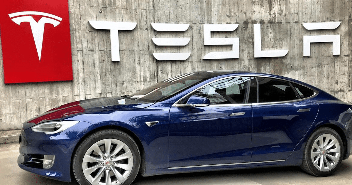 Automóvil Tesla azul aparcado frente a un muro de concreto con logo de Tesla en el fondo