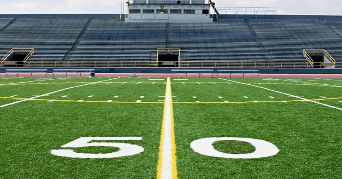 Campo de fútbol americano con césped artificial y el número 50 en el centro, gradas vacías al fondo en el estadio Kuykendall.