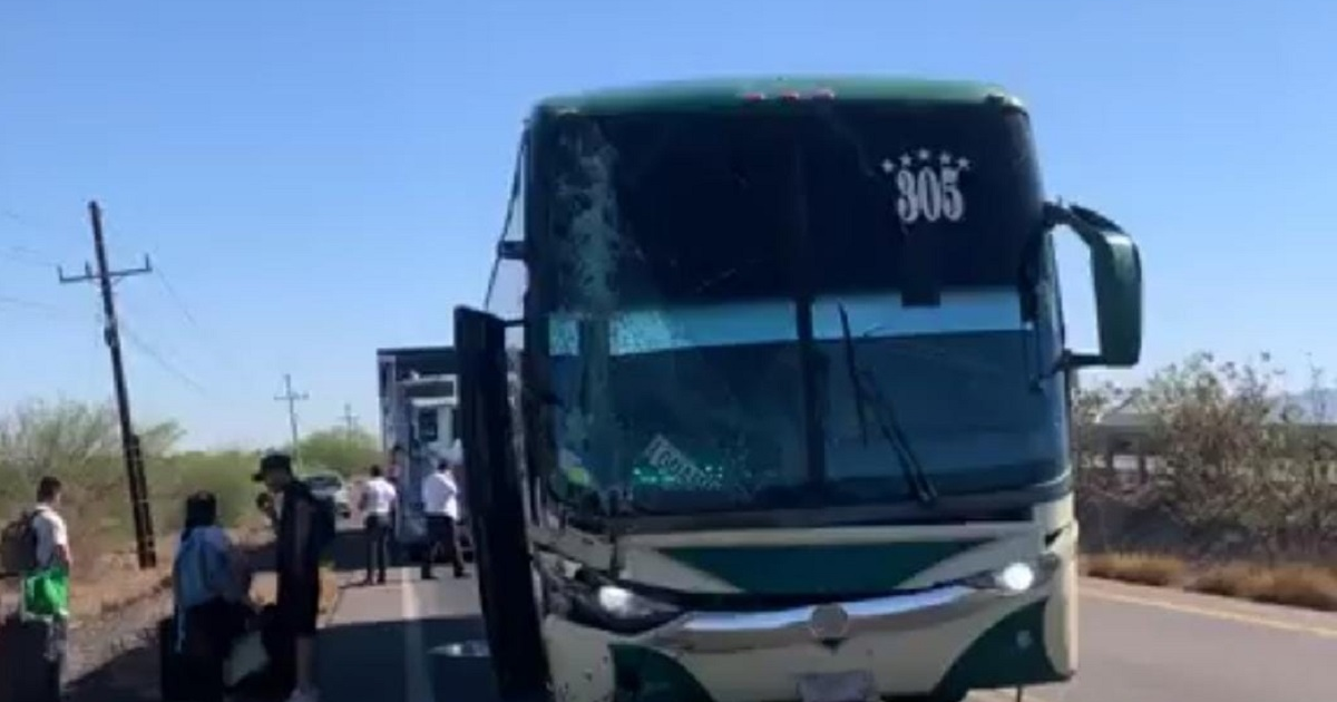 Autobús de pasajeros detenido en carretera desértica tras colisión, con cristal roto y pasajeros al costado.