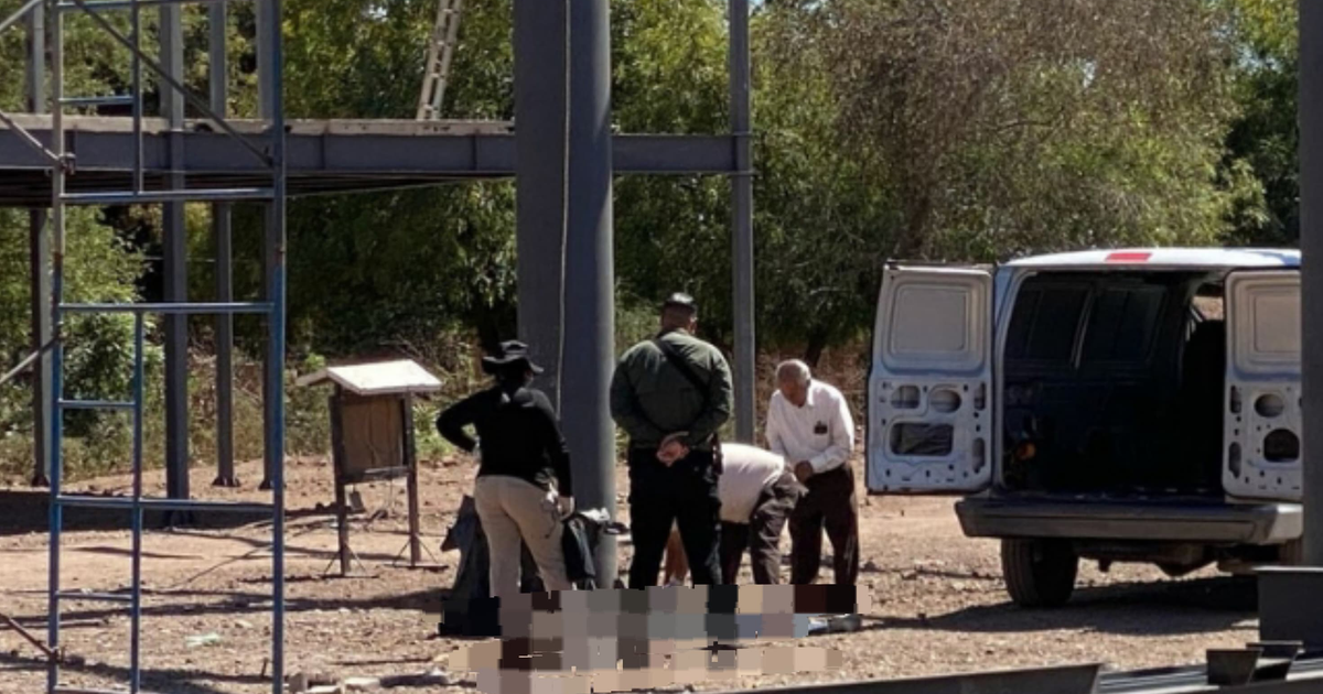 Grupo de personas en un sitio de construcción al aire libre, con estructura metálica y vehículo de transporte, relacionado con un accidente laboral fatal.