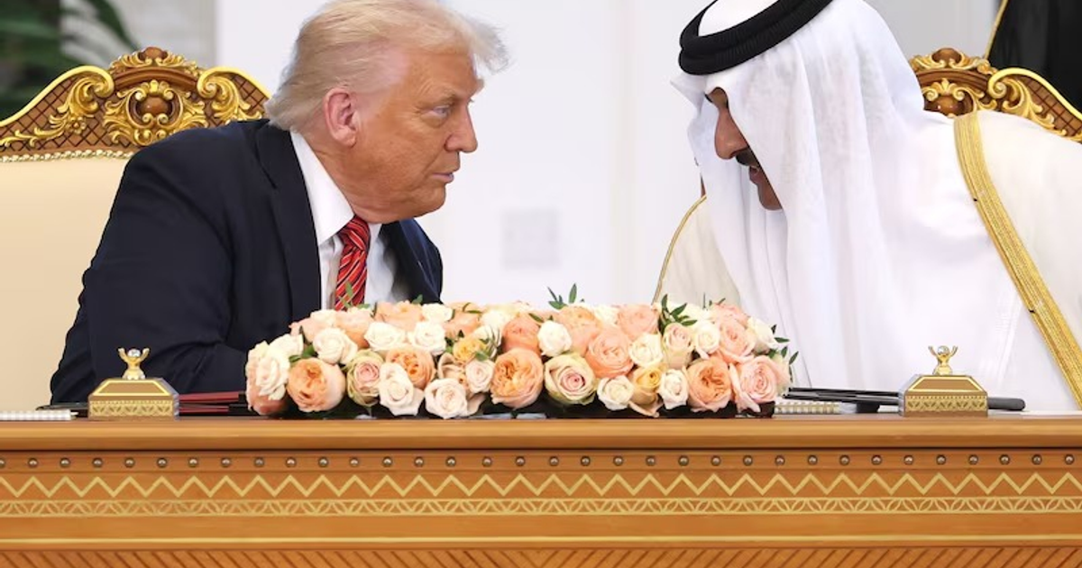 Dos hombres en conversación formal frente a un escritorio decorado con flores, en un contexto diplomático.