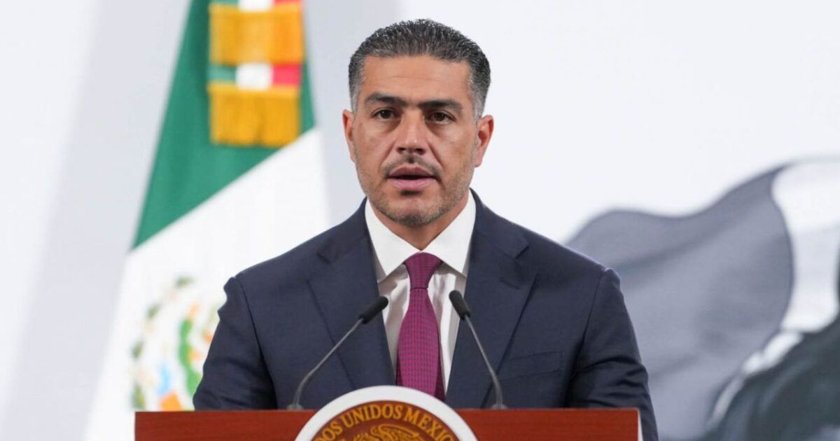 Hombre en podio con bandera mexicana, hablando sobre investigación de homicidios en CDMX.