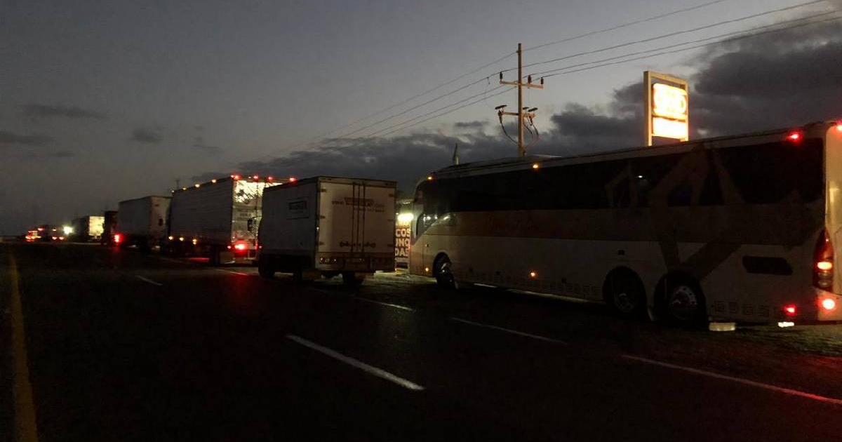 Fila de vehículos estacionados al anochecer en carretera Mazatlán-Culiacán, con camiones y autobús visibles, luces de vehículos y un letrero iluminado en la distancia.