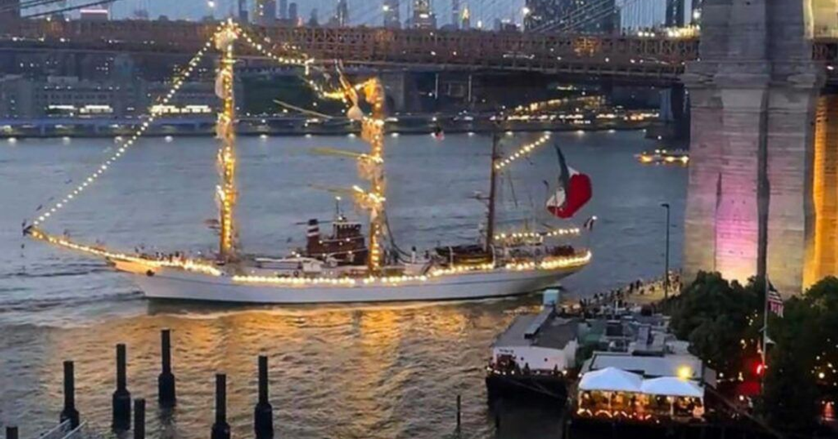 Barco iluminado con bandera de México navegando en río al amanecer, con puente y edificios al fondo.