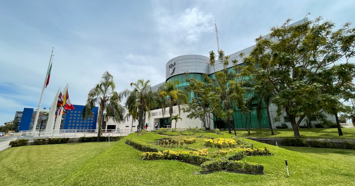 Edificio moderno de la UAS con fachada de vidrio, áreas verdes y banderas, incluyendo la de México, y un mural en una pared azul.