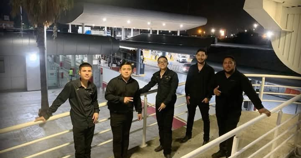 Cinco personas vestidas de negro posan en un entorno urbano nocturno con arquitectura moderna y luces en movimiento.