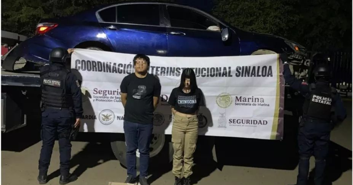 Dos personas detenidas frente a un banner de Coordinación Interinstitucional Sinaloa, con un vehículo azul y un agente de policía presente.