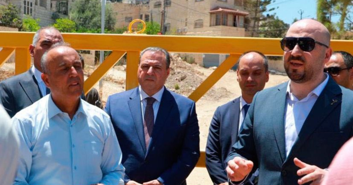 Grupo de diplomáticos en visita a Jenin, Cisjordania, con construcciones y vegetación de fondo.
