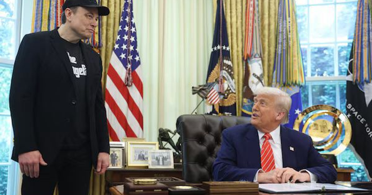 Dos hombres en una oficina oficial, uno de pie con chaqueta negra y otro sentado con traje azul y corbata roja, rodeados de banderas.