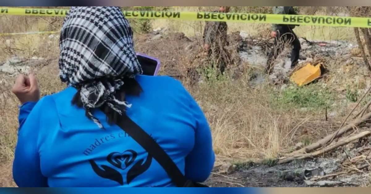 Persona de espaldas con pañuelo en la cabeza en un sitio de investigación de fosas clandestinas, rodeada de vegetación seca y cinta de precaución amarilla.