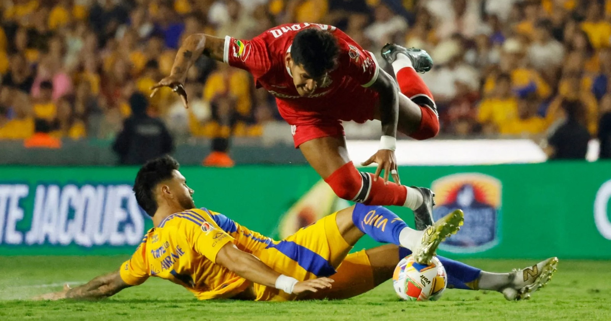 Jugador de camiseta roja en el aire tras choque con jugador de camiseta amarilla y azul en partido Tigres vs Toluca